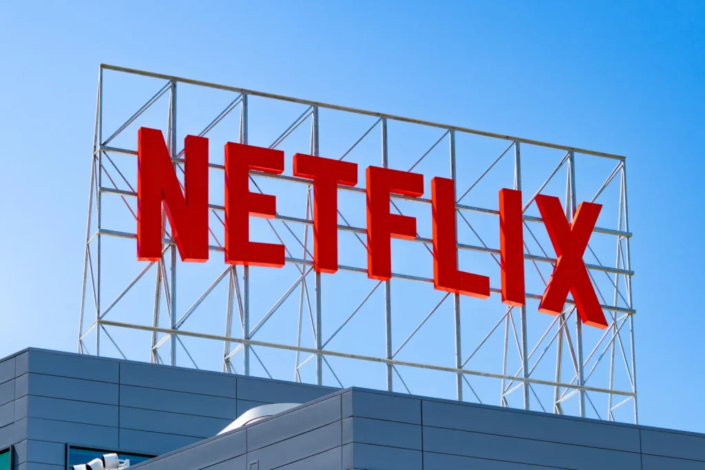 A low-angle view of large 3D red Netflix letters installed on a metal scaffolding frame atop a modern office building under a clear blue sky.